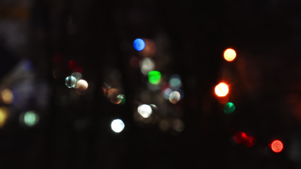 Timelapse of out of focus colorful traffic lights shimmering down a dimly lit street, creating a vibrant atmosphere in a calm neighborhood. The surrounding trees frame the mystical scene at night