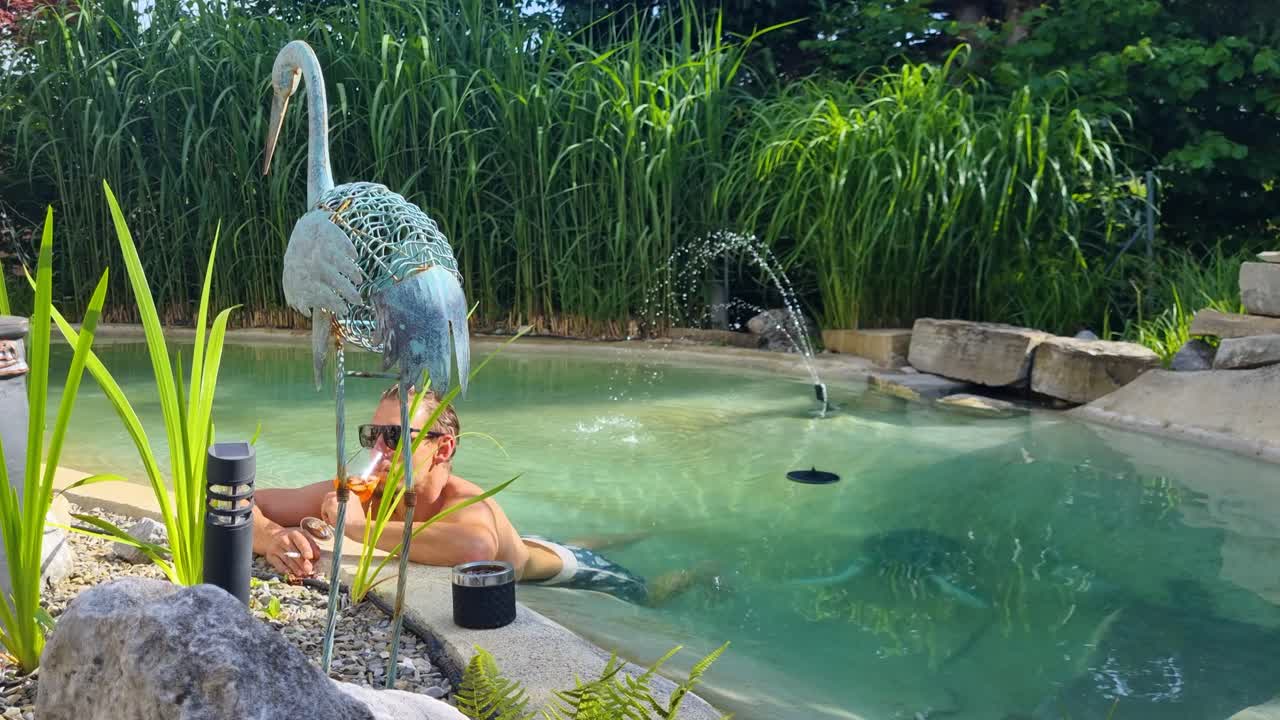Stylish man in sunglasses chills in a turquoise garden pool in Switzerland, sipping a cocktail and smoking, surrounded by reeds and tropical plants. Pure summer vibes on a hot day