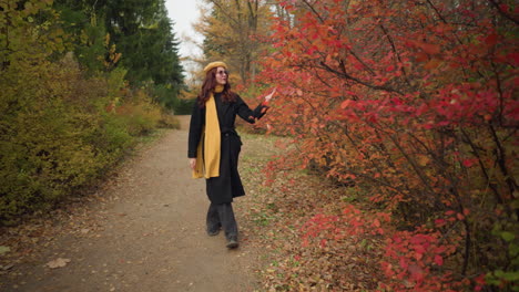 young lady in a yellow beret and muffler explores a woodland path, touching vibrant red autumn leaves, fully immersed in the serene ambiance