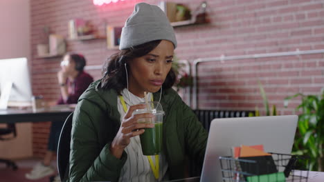 beautiful african american business woman using laptop computer typing browsing online messages drinking juice listening to music checking smartphone in modern trendy office