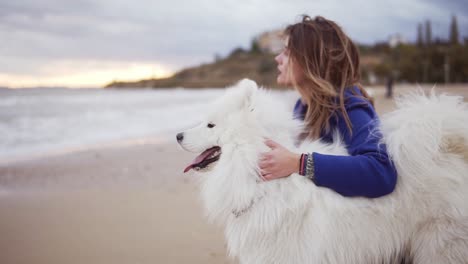 Side-view-of-a-young-woman-sitting-on-the-sand-and-embracing-her-dog-of-the-Samoyed-breed-by-the-sea.-White-fluffy-pet-on-the