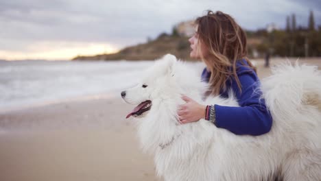 vista lateral de una mujer joven sentada en la arena y abrazando a su perro de la raza samoyed junto al mar. mascota peluda blanca en el