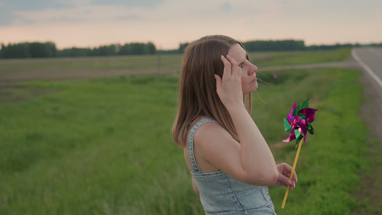 Lady adjusts hair while holding colourful pinwheel rotating in wind beside lush green grass field under cloudy sky conveying playful mood and gentle summer breeze