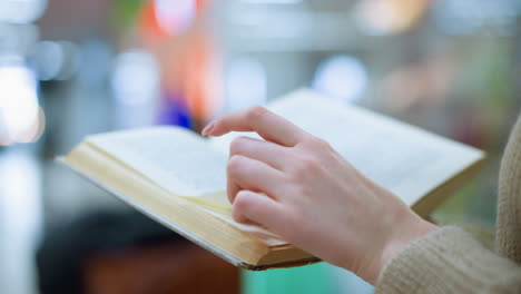 색 여성이 새 페이지로 돌아가는 가까운 면 뷰: 짜여진 금색 스웨터, 현대적인 구조, 흐릿한 그림과 초록색 장식 꽃, 근처에 쇼핑 가방