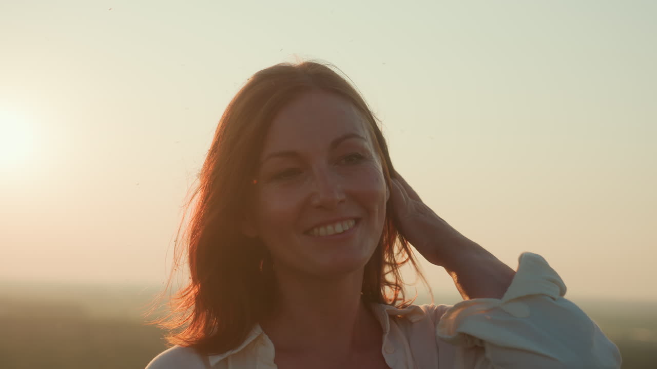 Close up young woman spinning joyfully on sunlit hilltop overlooking river valley and tall grass as breeze lifts hair and landscape blurs creating dreamy motion and freedom vibe in golden hour light