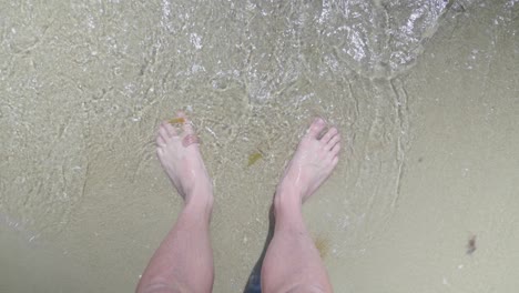 a point of view shot looking down at a mans feet while small waves lap at his toes