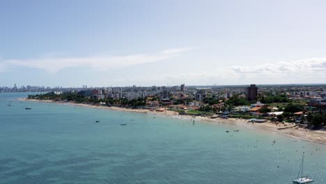 Aumento-De-Camiones-A-La-Derecha-Vista-Aérea-Extrema-De-La-Playa-Tropical-De-Bessa-En-La-Ciudad-Capital-De-Joao-Pessoa,-Paraiba,-Brasil-Con-Gente-Disfrutando-Del-Océano-Rodeada-De-Palmeras-Y-Edificios