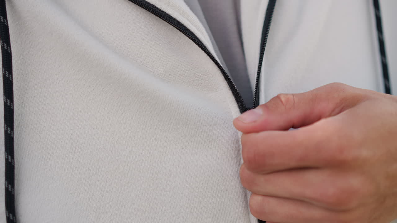 Primer plano de un hombre blanco subiéndose la cremallera de una sudadera con capucha, enfoque táctil en las manos y el tejido de punto texturizado, con detalle del cordón y costura a la vista, corredor preparándose para trotar y luego viajero asegurándose una capa, ambiente tranquilo y minimalista