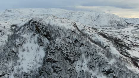 Drone-view-in-Tromso-area-in-winter-flying-over-a-snowy-mountain-peak-surrounded-by-the-ocean-in-Norway-and-wind-mills-on-the-horizon