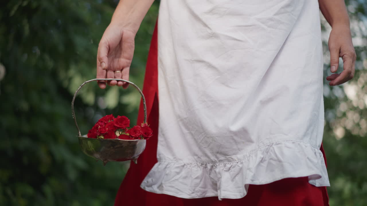 Primo piano di una donna bianca che tiene un cesto di petali rossi, con un grembiule bianco arricciato sopra una gonna rossa, mentre passeggia delicatamente attraverso un giardino rustico lussureggiante, sotto una morbida luce solare con effetto bokeh, con un orlo di pizzo tattile, in un'atmosfera rustica e romantica.