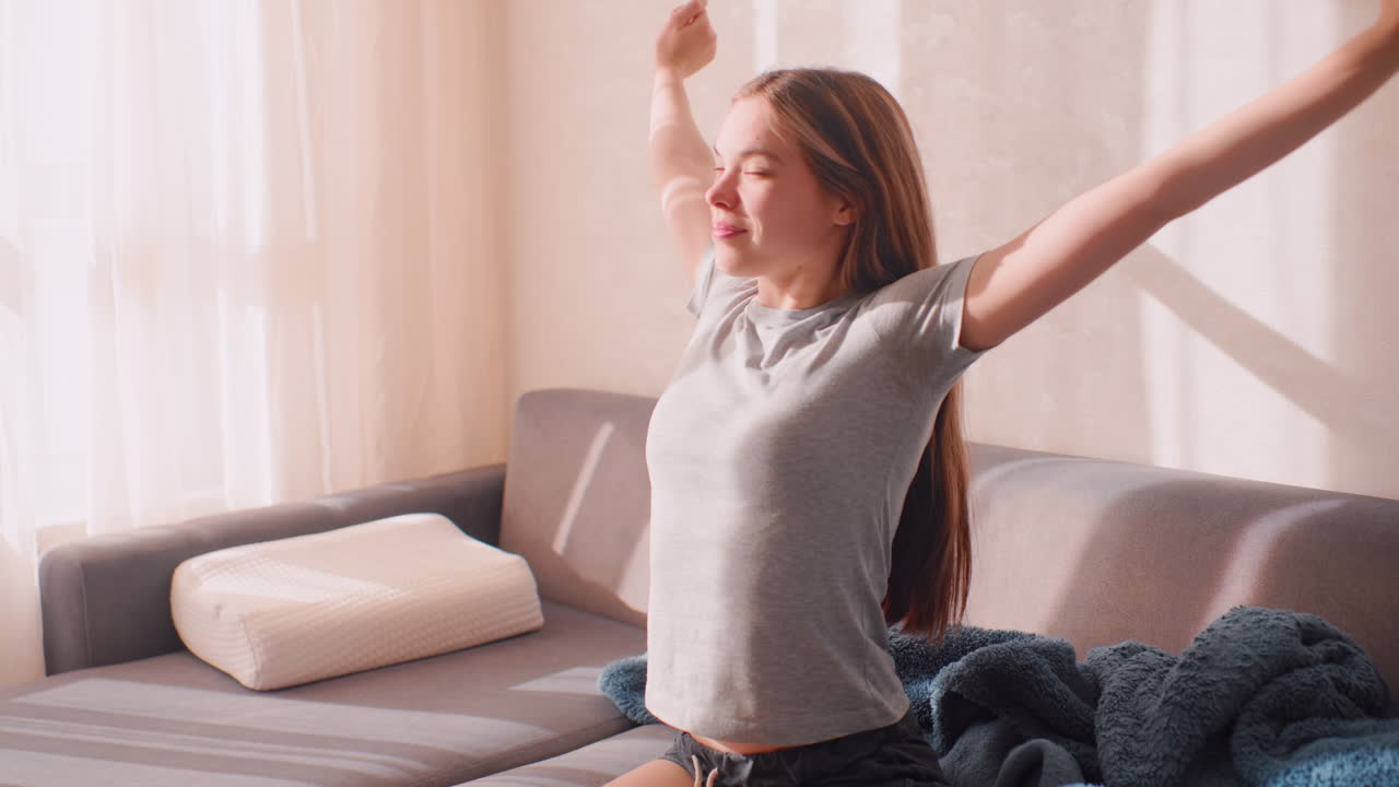 Young woman wakes up sitting on couch with eyes closed stretching shoulders enjoying warm morning sunlight through window with cozy blanket beside her
