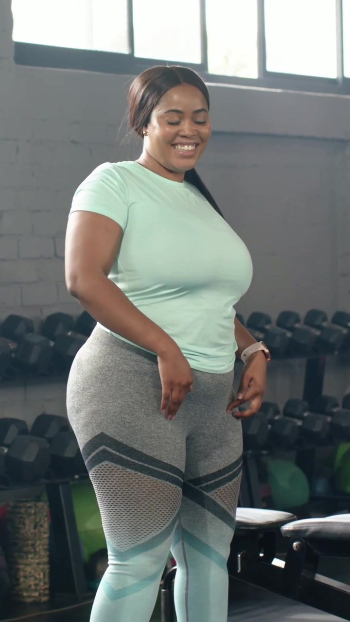 Video verticale: Donna che ride con maglietta color menta vicino ai pesi in palestra, sistema i fianchi reagendo a un segnale fuori campo.