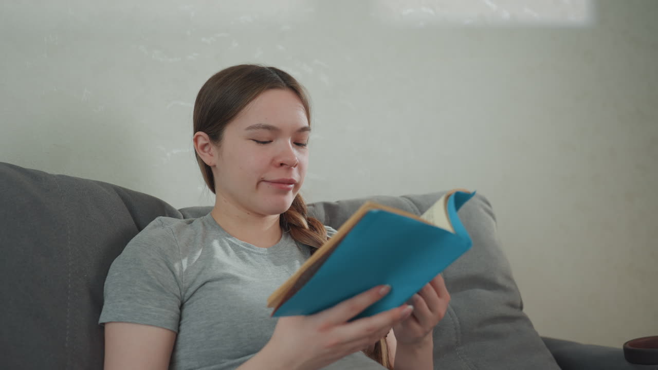 Pregnant woman reading novel in quiet living room, blue paperback open, relaxed posture against cushions, braided hair, reflective expression, evening calm, gentle breathing, mindful rest