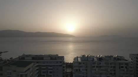 Drone-shot-above-the-city-Vlore-Albania-looking-over-the-buildings-and-harbour-with-ferry-with-the-sea-and-beach-underneath-during-sunset-in-the-evening-with-an-orange-glow-LOG