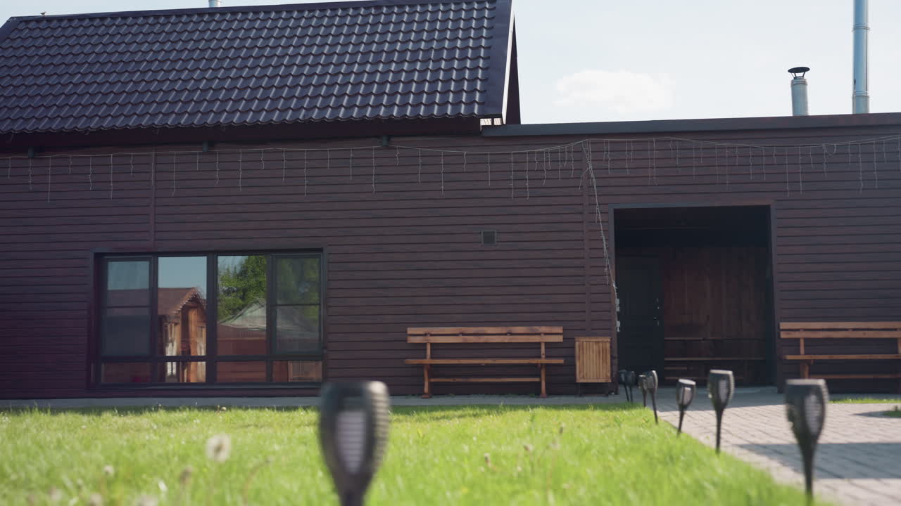Brown wooden house in sunny fenced compound featuring paved walkway leading to entrance, bench seating along exterior wall, visible wooden swing structure, small cabin reflection in window