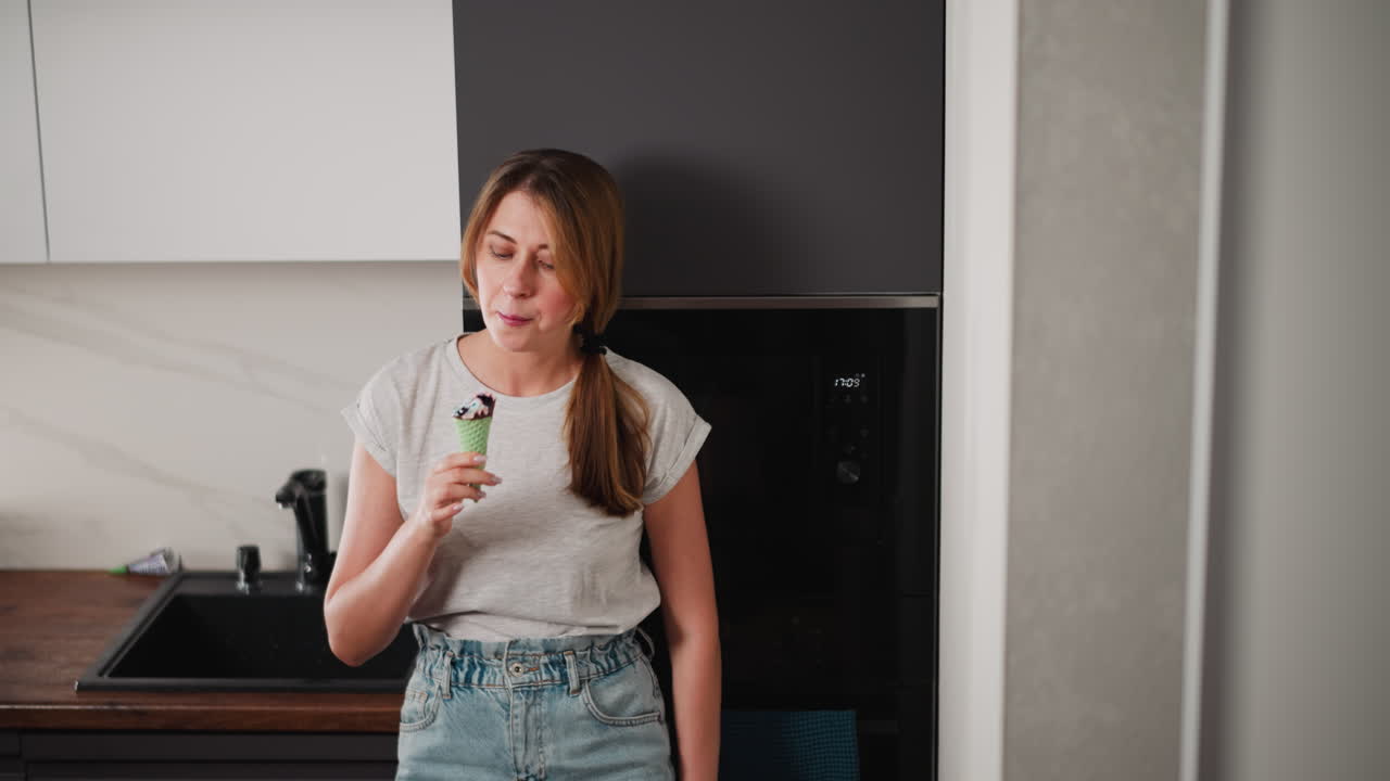 Elegant lady wearing grey t-shirt and blue jeans enjoys ice cream with green cone, standing casually near stylish modern kitchen setup with black fridge and white cabinets, savoring sweet treat