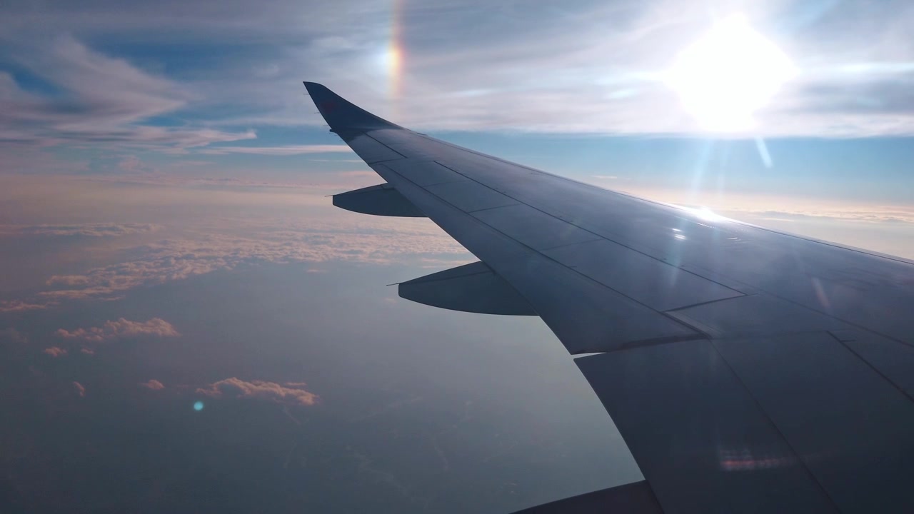 Raro fenomeno ottico del parelio nell'alta atmosfera, fotografato da un finestrino passeggero, con l'ala dell'aereo sagomata contro un sole luminoso e basso all'orizzonte e una nuvola morbida dai colori pastello.