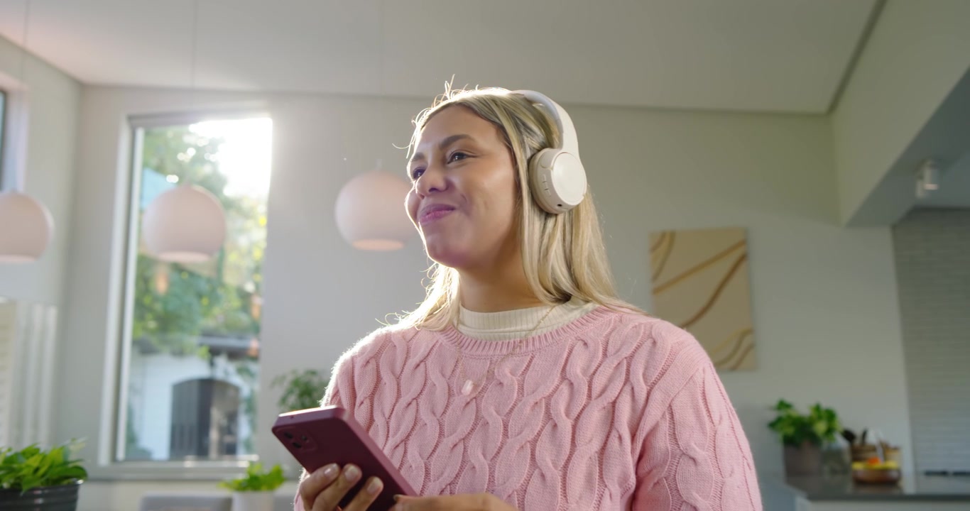 Donna afroamericana di mezza età che tocca le cuffie e scorre il telefono in cucina mentre ascolta un podcast.