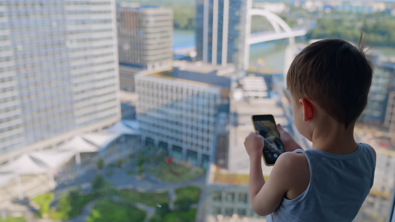 Child plays on phone by window. A young boy stands at a high-rise window looking out at the city while using his smartphone in the evening
