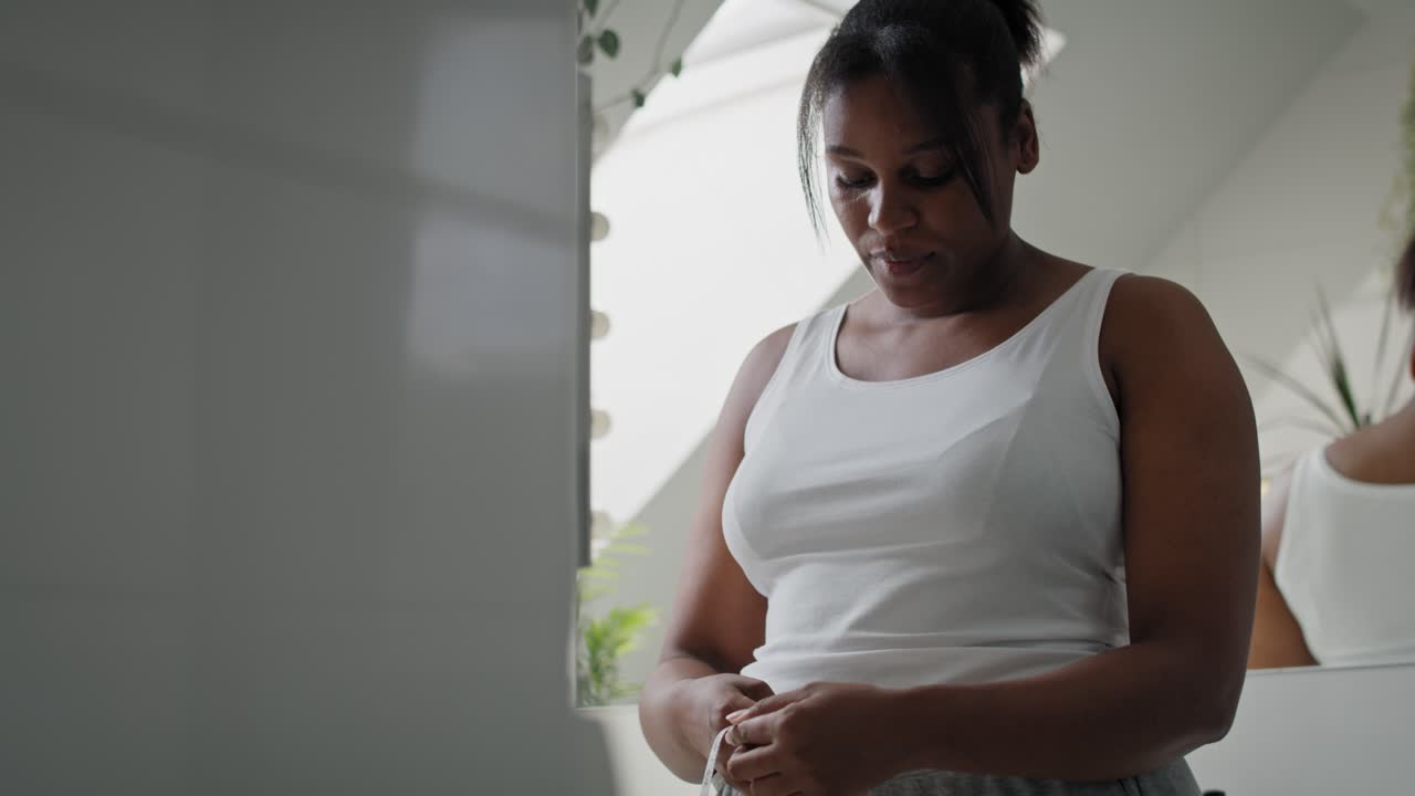 Donna afroamericana insoddisfatta in piedi in bagno che si misura la vita.