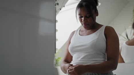 Dissatisfied-African-American-woman-standing-in-the-bathroom-and-taking-measures-on-her-waist.