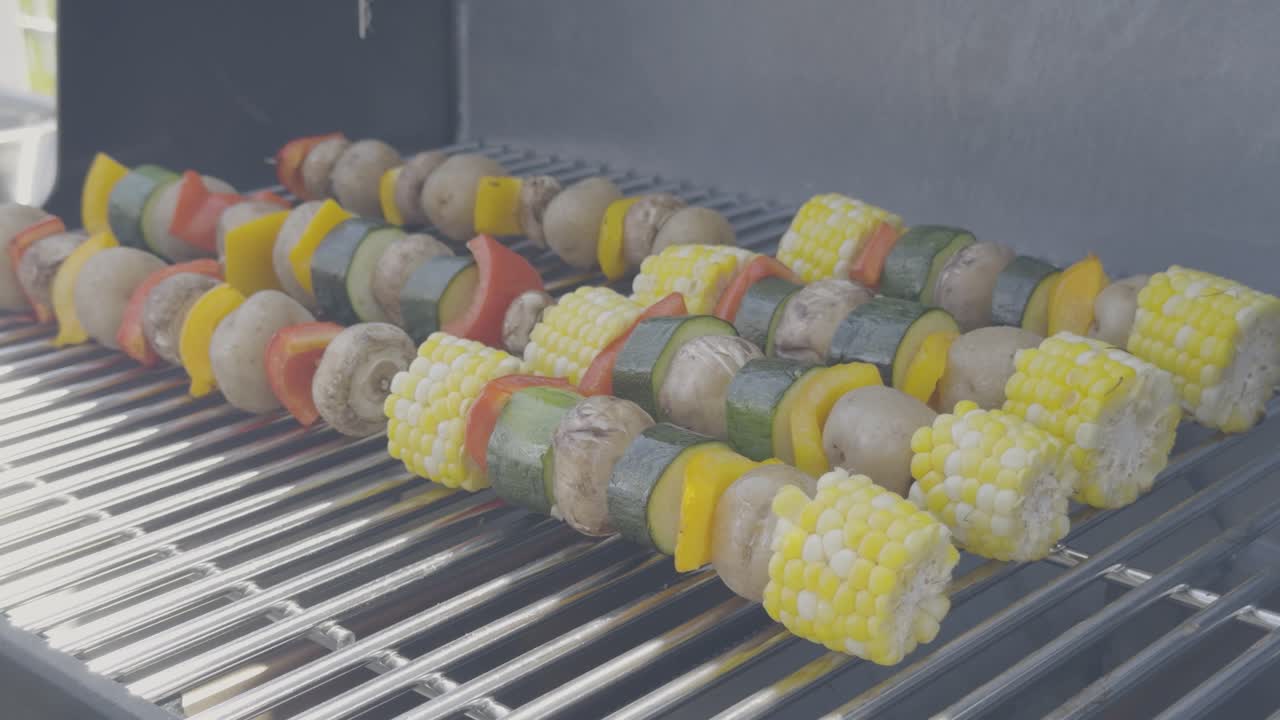 Side angle ofvegetable skewers with corn, pepper, zucchini, potatoes and mushrooms are being cooked on a stainless steel bbq and brushed with oil. Smoke and flames rise from the grill