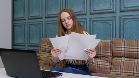 Woman-sittingat-home-looking-at-documents-analyzes-data-checks-papers-working-on-laptop-computer