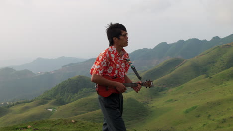 close up shot of an asian man standing on top on a mountain singing while playing ukulele on a windy day