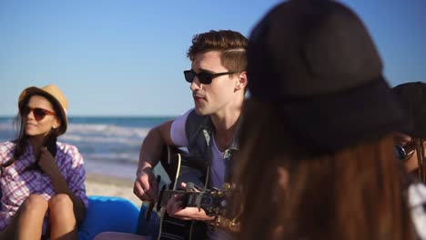 Young-man-playing-guitar-among-group-of-friends-sitting-on-easychairs-on-the-beach-and-singing-on-a-summer-evening.-Slowmotion-shot