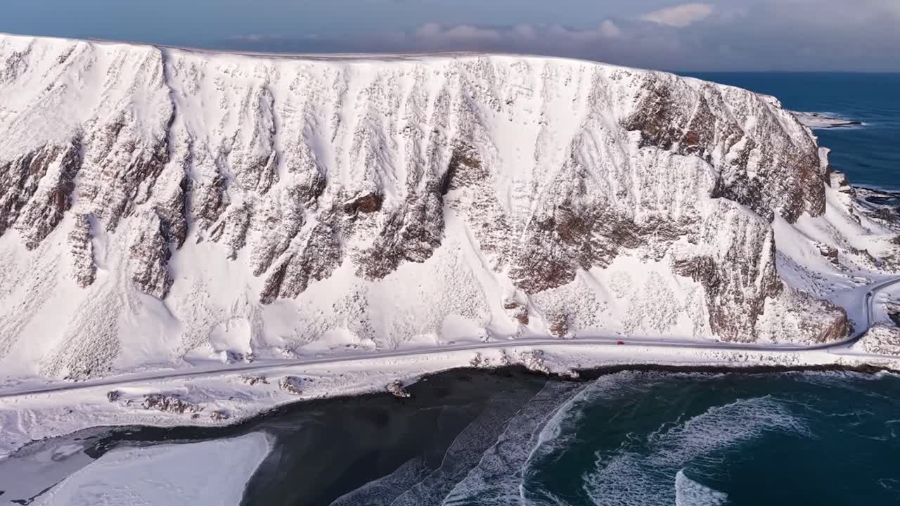 Aerial drone footage of a Norwegian shoreline with mountain and a road during winter time while sun is shining. The road and mountain is on the coast of ocean blue sea water that has big waves.