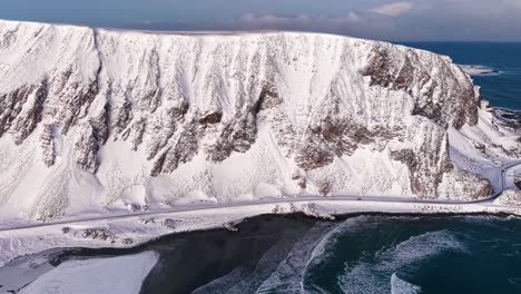 aerial drone footage of a norwegian shoreline with mountain and a road during winter time while sun is shining. the road and mountain is on the coast of ocean blue sea water that has big waves.