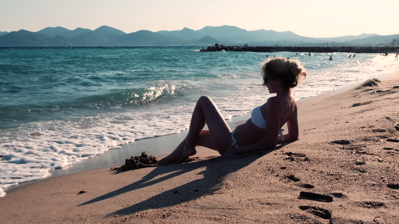 A young woman in a white bikini enjoys the sun on a sandy beach while a cruise ship sails in the distance