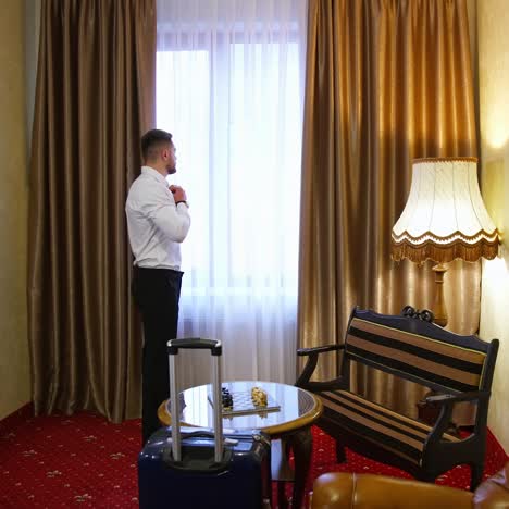 young business man in a hotel room. man in elegant clothing standing near the window. traveler arrived to a hotel with suitcase. business journey