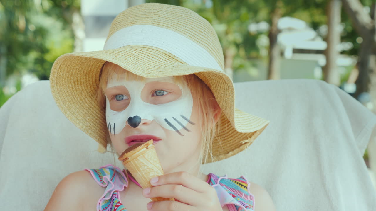 그녀의 얼굴에 아쿠아 그림을 가진 소녀는 어린 개념으로 리조트에서 아이스크림을 먹습니다.