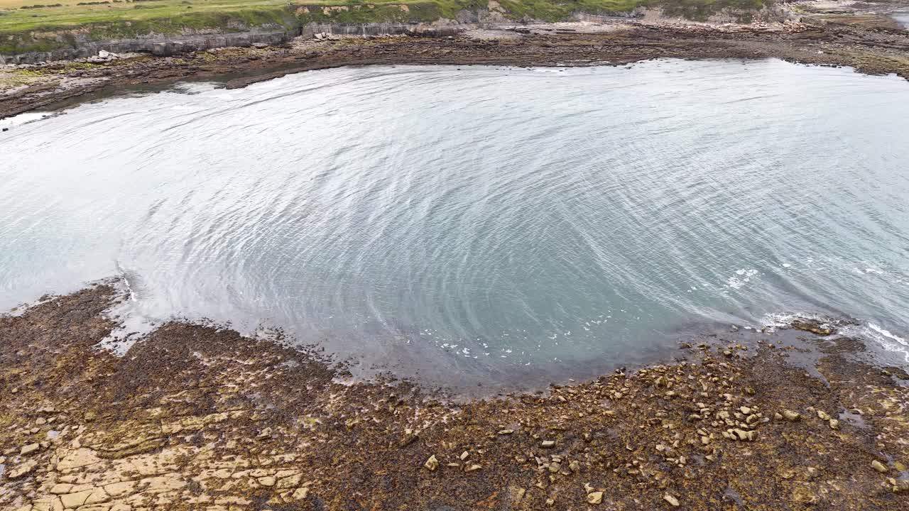Drone camera glides above a rugged rocky shoreline and tidal pool under overcast daylight, revealing textured rock formations and gentle water movement along the coast