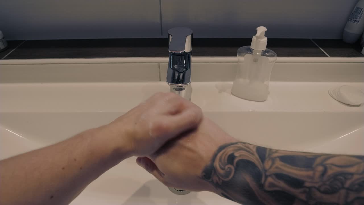 Førstepersonsoptagelse af en ung mand med tatoveringer på armen, der vasker hænder med sæbe under rindende vand på et moderne badeværelse