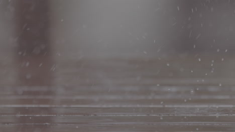 Close-up-shot-of-rain-drops-falling-on-a-wooden-table-making-splashes-with-chairs-in-the-background-in-slowmotion-LOG