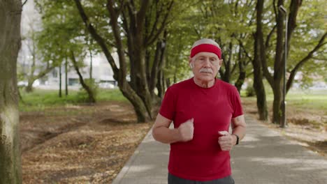 Male-senior-person-running-along-the-road-in-park.-Mature-runner-man-training,-listening-music