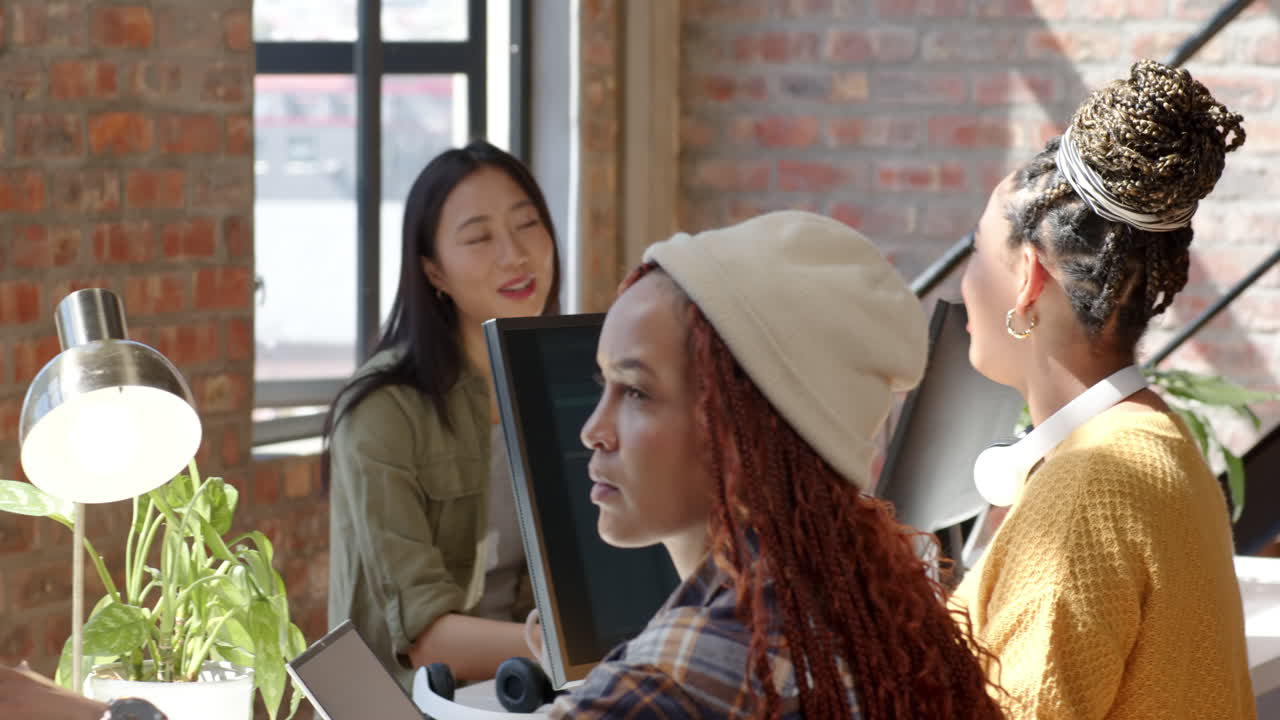 Diverse female team collaborating in modern office, discussing coding project with computers