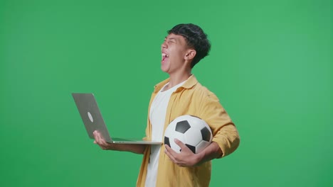 vista lateral de un niño asiático con una pelota gritando gol alegrándose por el equipo ganador mientras anima el fútbol en una computadora portátil en el estudio de pantalla verde