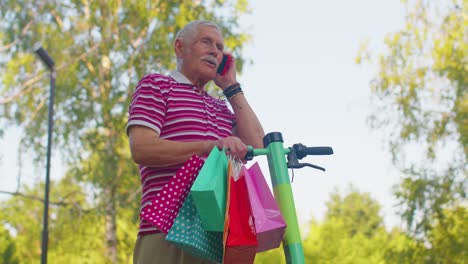 anciano abuelo hombre en scooter eléctrico con bolsas de compras de colores hablando en el teléfono móvil