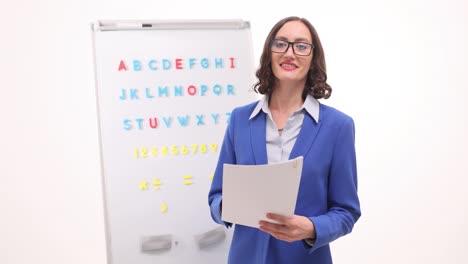 smiling woman teacher holding papers in front of an educational whiteboard