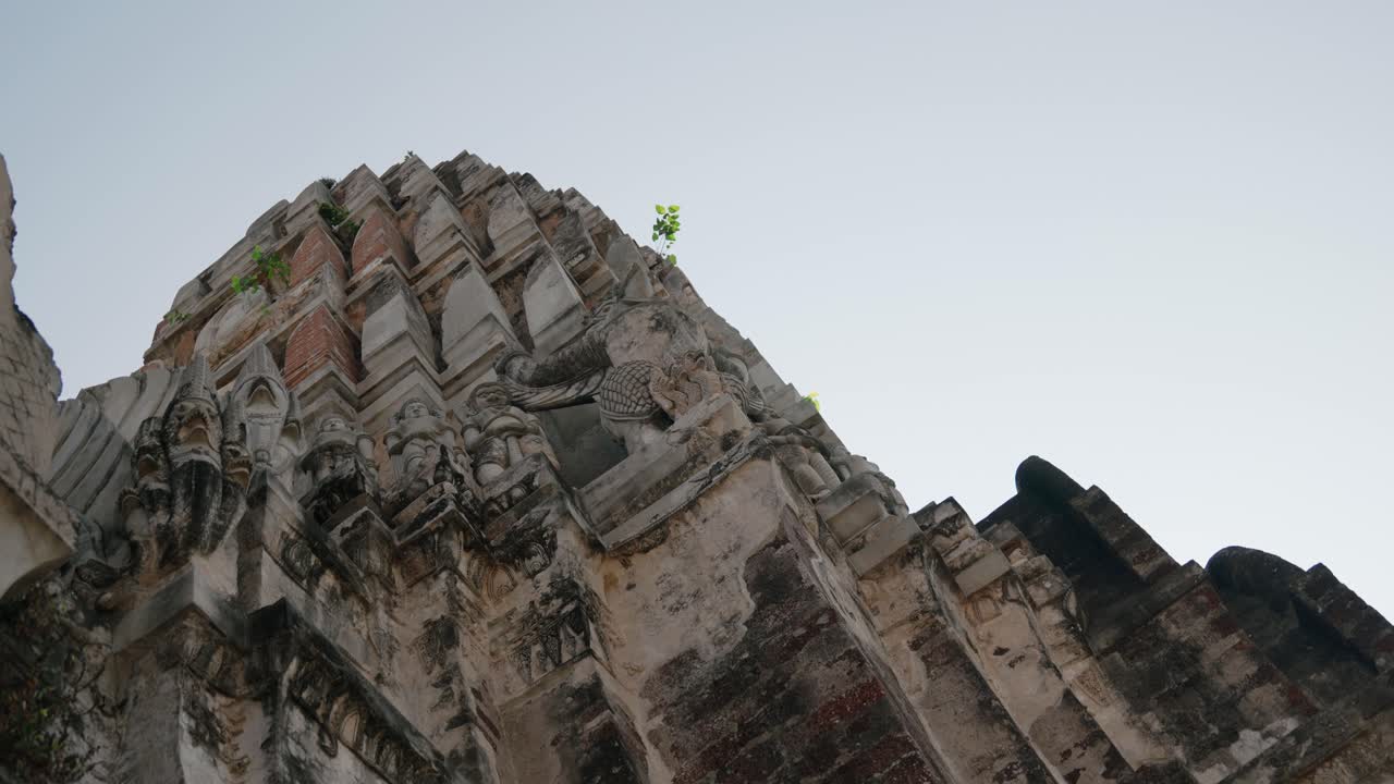 Ancient stone temple structure in Ayutthaya with intricate carvings and weathered details, captured from a low angle against the clear sky