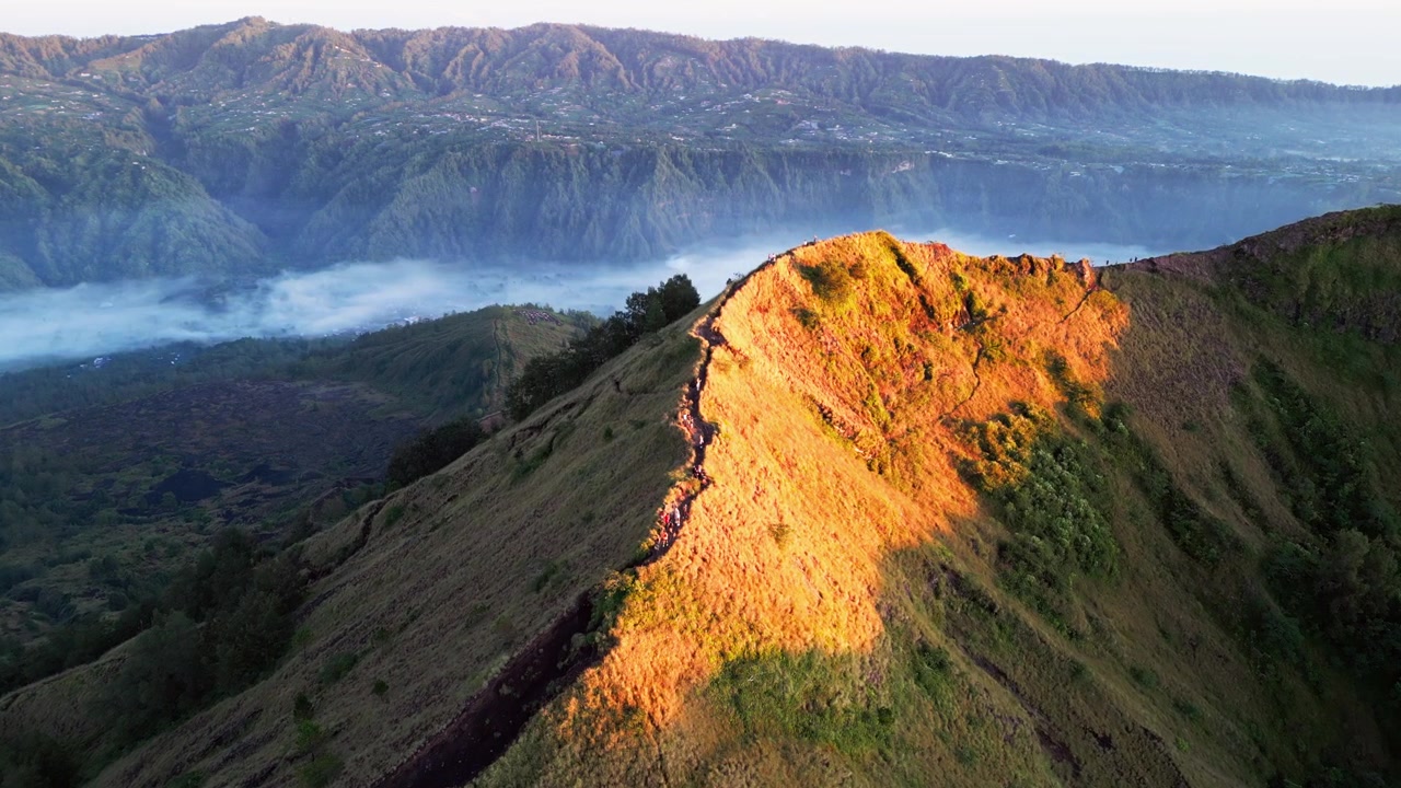 Escursionisti che camminano lungo una cresta montuosa illuminata dal sole all'alba sopra una valle vulcanica nebbiosa e un drammatico paesaggio montano a Bali, riprese aeree con drone in avvicinamento.