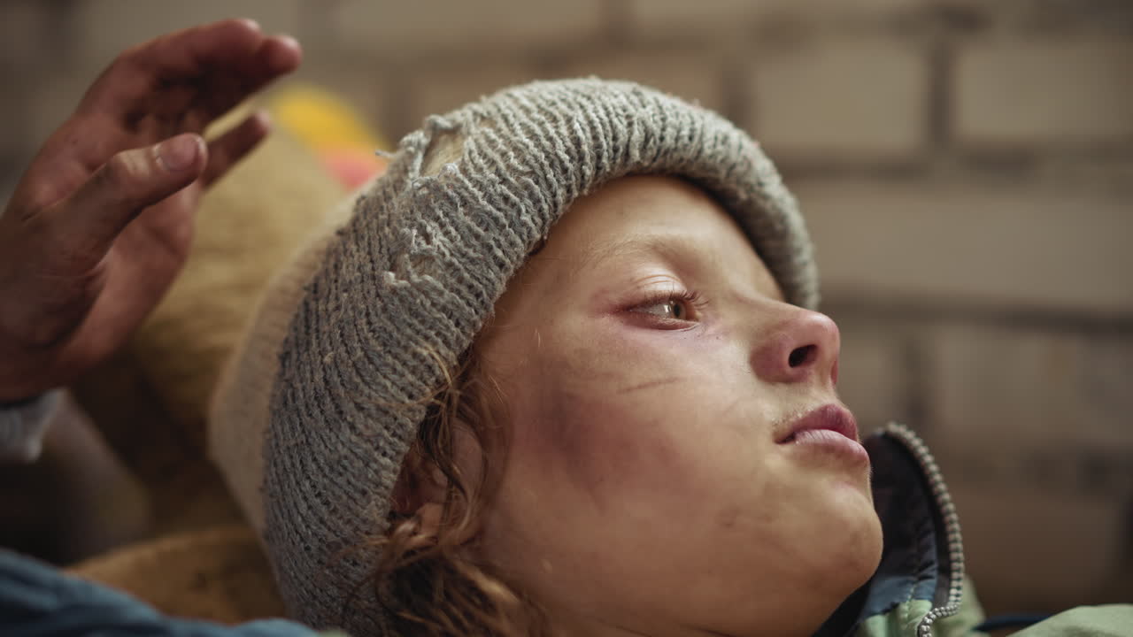 Close up of child face lying sideways wearing worn knit hat with bruises and dirt, while hand gently taps head showing concern, compassion, and human connection in difficult situation indoors