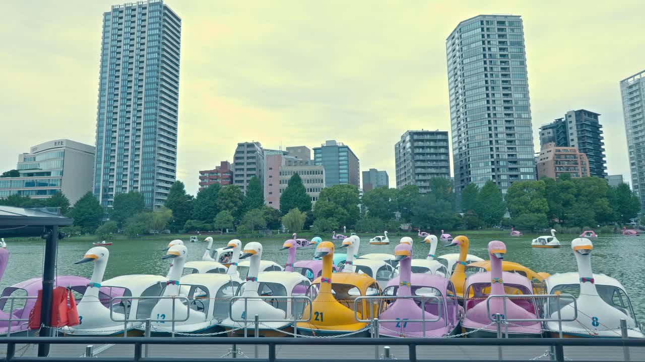 Colorful swan boats float on Shinobazu Pond in Ueno Park, with Tokyo's modern skyline visible in the background, blending nature and urban life.