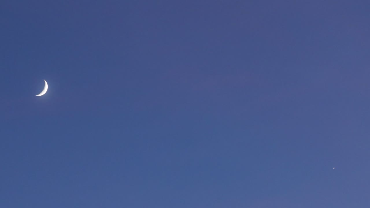 "Drone shot at 167mm of a crescent moon and star rising in a clear blue sky at dusk, both ascending on opposite sides of the frame with no other elements in the calm, peaceful sky.