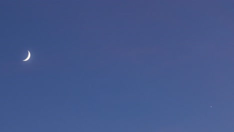 "drone shot at 167mm of a crescent moon and star rising in a clear blue sky at dusk, both ascending on opposite sides of the frame with no other elements in the calm, peaceful sky.