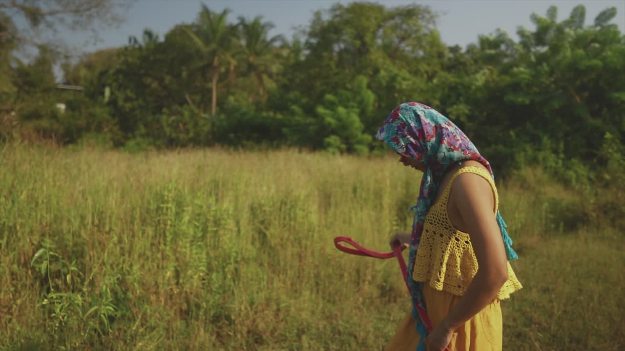 una hermosa joven vestida de amarillo y bufanda floral camina por los campos con un cinturón en las manos y gafas de sol