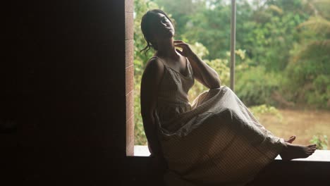joven sentada pensativa junto a una ventana en un entorno rústico, reflejando en silencio, con exuberante vegetación afuera
